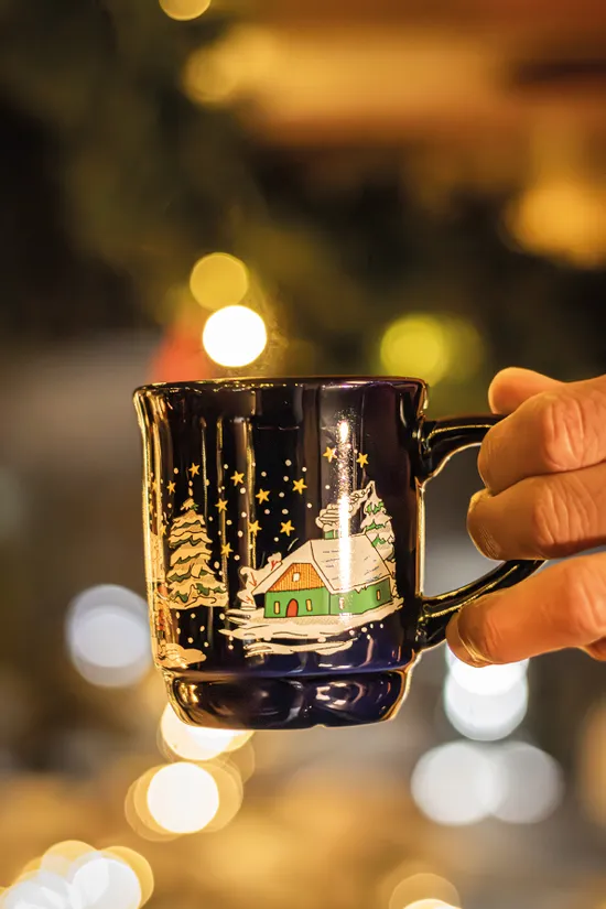 Hand holding festive Christmas mug with snowy village design and warm bokeh lights